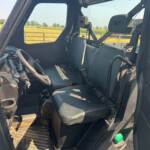 Interior of a truck cab with steering wheel, bench seat, and instrument panel; open door reveals a rural field outside.