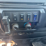 Dashboard panel with four vertical switches: electronic parking brake, a switch with an orange indicator, a differential/axle switch, and a windshield wiper/washer switch with a blue light; climate knobs below.