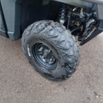 Close-up of a rugged off-road tire and suspension on a green utility vehicle, dirt and gravel on the ground nearby.
