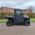 Side view of a dark rugged off-road utility vehicle with large tires parked on a driveway, hedges in front of red-tiled houses.