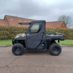 Black Polaris Ranger utility vehicle parked on a street with hedges and red-tiled houses in the background under a cloudy sky.