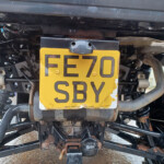 Rear view of a motorcycle showing a yellow license plate mounted on the back, with exposed frame, suspension, and mechanical parts surrounding it.