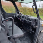Interior of a rugged off-road vehicle showing a steering wheel, dashboard, pedals, and a large treaded tire inside a wet cabin. Outside visible are raindrops on windows and a rural yard scene there.