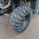 Close-up of an off-road vehicle's front wheel with a large knobby tire and exposed suspension, showing some rust on nearby exhaust components.