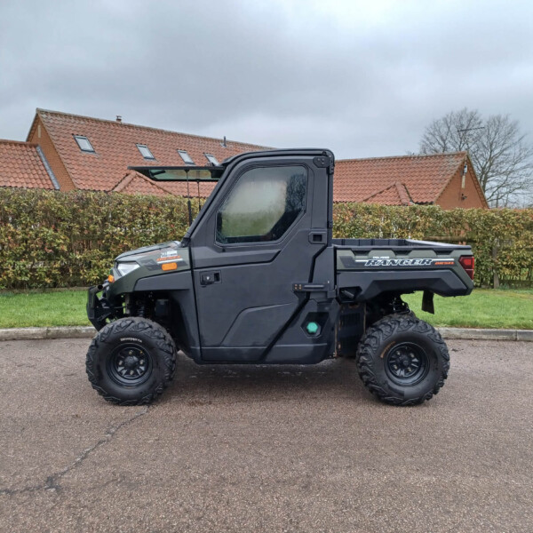 Black Polaris Ranger utility vehicle parked on a street with hedges and red-tiled houses in the background under a cloudy sky.