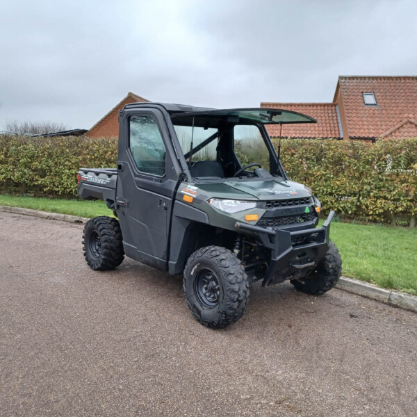 Dark gray off-road utility vehicle with open cabin parked beside a hedge and red-tiled houses in a suburban street under a cloudy sky.