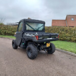 Rear three-quarter view of a black Polaris Ranger off-road utility vehicle parked on a residential street, with hedges and red-tiled houses in the background.