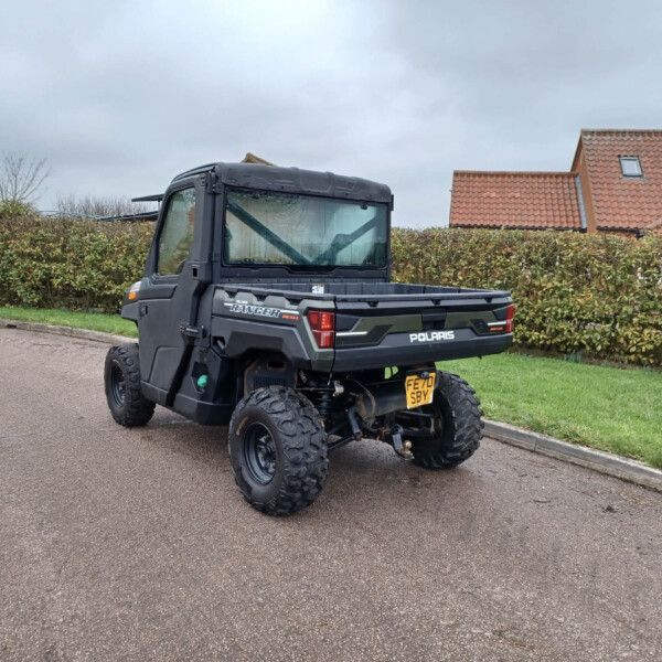Rear three-quarter view of a black Polaris Ranger off-road utility vehicle parked on a residential street, with hedges and red-tiled houses in the background.