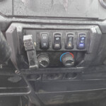 Close-up of a dusty vehicle dashboard showing a row of five black control buttons with various symbols (parking brake, other vehicle controls) and a blue-illuminated windshield wiper icon on the right.