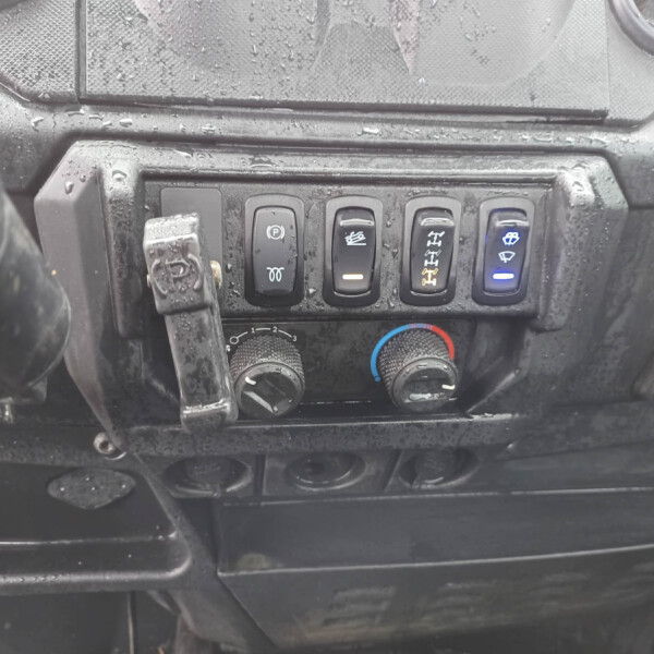 Close-up of a dusty vehicle dashboard showing a row of five black control buttons with various symbols (parking brake, other vehicle controls) and a blue-illuminated windshield wiper icon on the right.