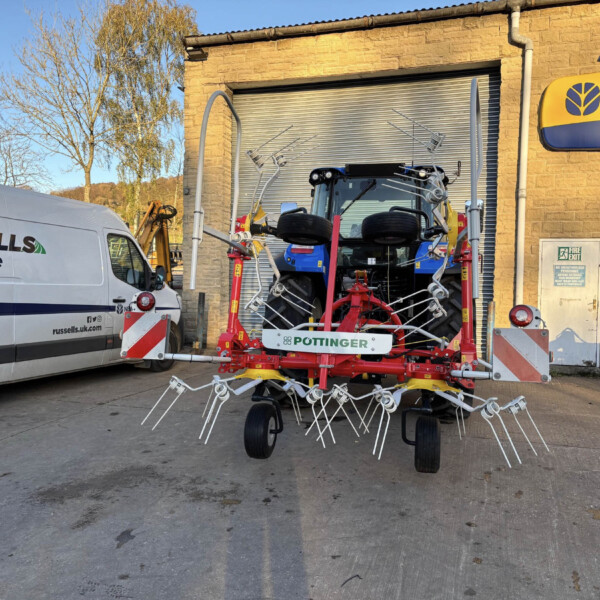 Industrial yard with red multi-row farming implement and a blue tractor, beside a Russells Agriculture van outside a brick building with a New Holland sign on the wall.