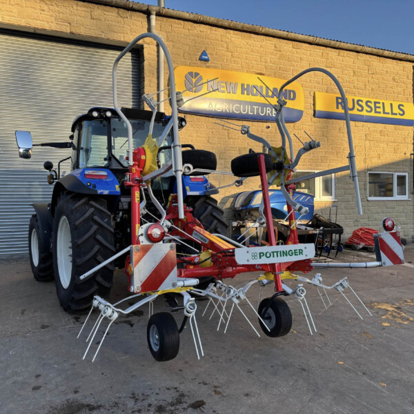 Blue New Holland tractor with multiple attached farming implements on a display yard in front of a brick building with New Holland Agriculture and Russell's signage.