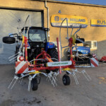 Large agricultural implement with many tines and a red frame parked outside a New Holland dealership sign.