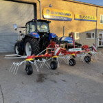 Blue New Holland tractor parked outside a farm equipment shop with yellow signage reading 'New Holland Agriculture' on a brick building.