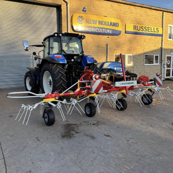 Blue New Holland tractor parked outside a farm equipment shop with yellow signage reading 'New Holland Agriculture' on a brick building.