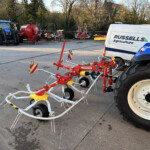 Blue tractor and red farming implements parked in a yard near a Russell's Agriculture van in the background.