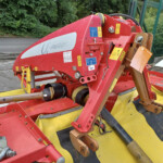 Red agricultural implement with hydraulic arms and yellow safety panels, parked outdoors near a fence and greenery, showing warning stickers and control levers.