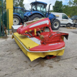 Red Novacat 301 mower attached to a trailer in an outdoor equipment yard, with blue tractors and a white van in the background.