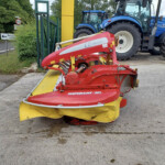 Red mowing attachment labeled NOVACAT 301 in the foreground, with a blue tractor and a road sign in the background.