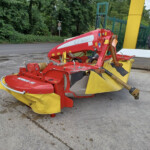Red farm implement with yellow guards parked on a paved surface beside a green fence and tractor in the background, outdoors near trees.