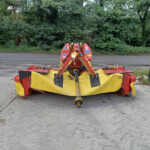 Red farming implement with a yellow cover sitting on a paved area, green trees in the background