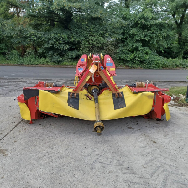Red farming implement with a yellow cover sitting on a paved area, green trees in the background