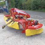 Red agricultural mower attachment with a yellow protective skirt parked on a concrete area beside a street, with a blue fence and green trees in the background.