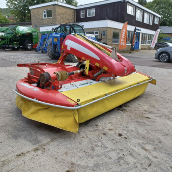 Red front-mounted mower with yellow guards in a farm equipment lot, green tractors in the background.