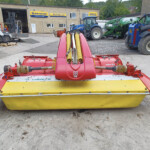Close-up of a red and yellow farming mower with yellow rollers in a tractor yard workshop scene behind it.