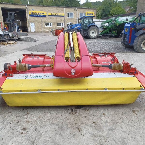 Close-up of a red and yellow farming mower with yellow rollers in a tractor yard workshop scene behind it.