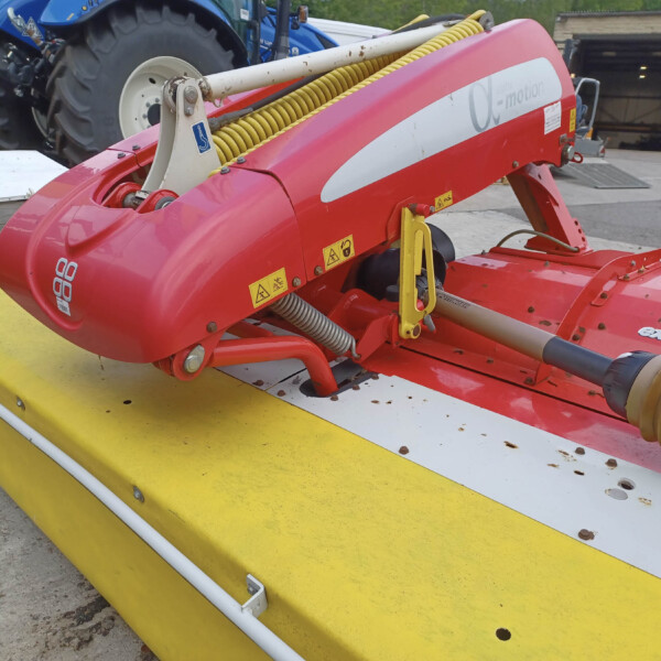 Close-up of a red agricultural machine with a yellow spiral hose, mounted on a yellow platform; tractors and a farm building visible in the background.