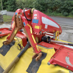 Red and yellow farming implement with hydraulic arms parked on a roadway, greenery in the background.