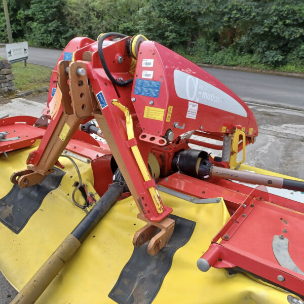 Red and yellow farming implement with hydraulic arms parked on a roadway, greenery in the background.