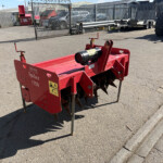 Red agricultural spiker attachment labeled 'Spiker 1350' standing on legs in a paved yard with fencing and parked vehicles in the background.