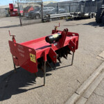 Red agricultural spike harrow/soil aerator labeled 'Spiker 1350' standing on metal legs in a paved outdoor lot with a fence and parked vehicles in the background