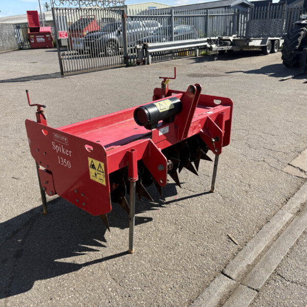 Red agricultural spike harrow/soil aerator labeled 'Spiker 1350' standing on metal legs in a paved outdoor lot with a fence and parked vehicles in the background