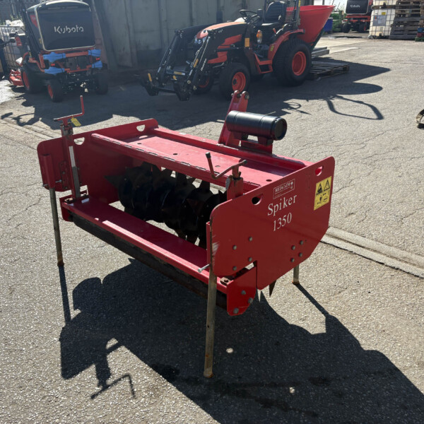 Red agricultural tiller attachment labeled 'Spiker 1350' in a yard with parked tractors and pallets in the background.