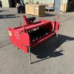 Red agricultural implement labeled “Spiker 1350” with spiked rollers, standing on legs in a concrete yard near a warehouse and pallets in the background