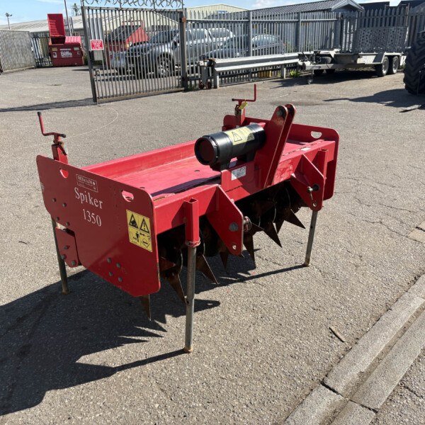 Red agricultural spiker attachment labeled 'Spiker 1350' standing on legs in a paved yard with fencing and parked vehicles in the background.