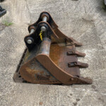 Rusty metal loader attachment resting on a rough concrete surface, with a yellow tag attached.
