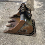 Close-up view of a rusted excavator bucket attachment resting on a rough concrete surface, with a fence visible in the background on the right.