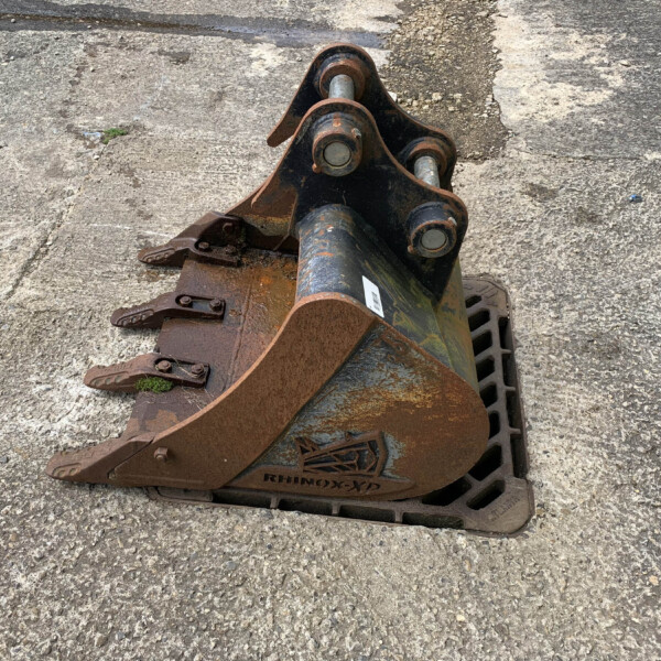 Close-up view of a rusted excavator bucket attachment resting on a rough concrete surface, with a fence visible in the background on the right.