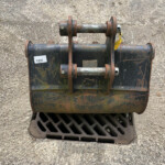 Rusty excavator bucket attachment with bolts and pins resting on a dirty black bucket, on a rough concrete surface beyond a drainage grate.