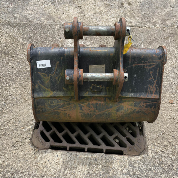 Rusty excavator bucket attachment with bolts and pins resting on a dirty black bucket, on a rough concrete surface beyond a drainage grate.