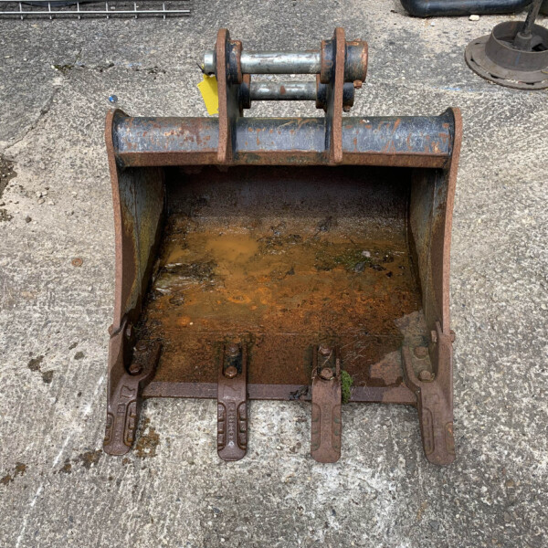 Rusty metal industrial bucket attachment with a hinged frame and hydraulic pins, sitting on a rough concrete surface with muddy water inside the bucket.