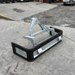Metal trailer frame with a black rubber bumper resting on a concrete surface in an industrial yard with trucks in the background.