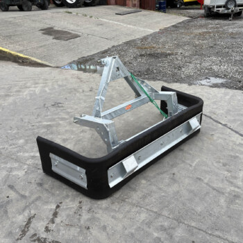 Metal trailer frame with a black rubber bumper resting on a concrete surface in an industrial yard with trucks in the background.