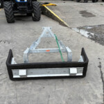 Close-up of a metal trailer frame with a black bumper on a concrete yard; large tires and machinery visible in the background.