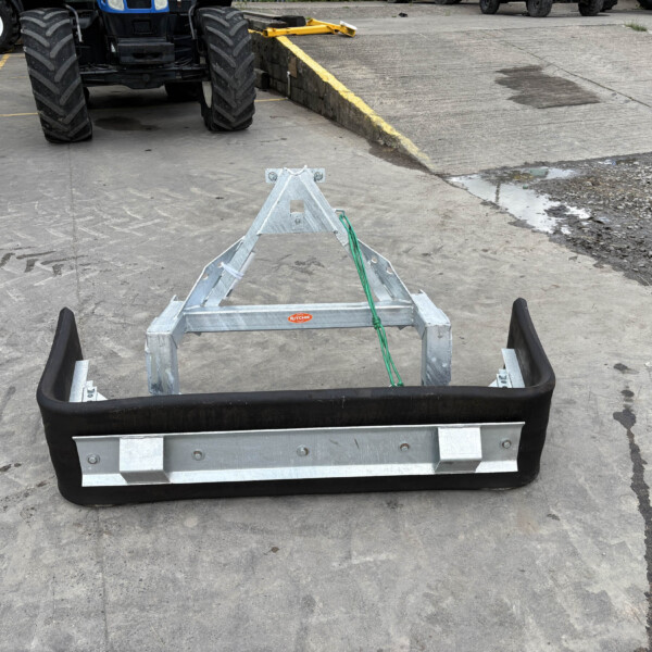 Close-up of a metal trailer frame with a black bumper on a concrete yard; large tires and machinery visible in the background.