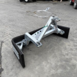 Metal agricultural hitch/attachment on a concrete surface, with a hydraulic-like arm and bolts, tractors visible in the background